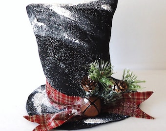 May include: A black top hat decorated for winter. The hat is dusted with white glitter and has a red plaid ribbon around the base. A sprig of pine, two pinecones, and a small bell are attached to the ribbon.