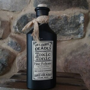 May include: A black glass bottle with a cork stopper and a brown twine bow. The bottle has a label that reads "DRS DIRE & DEADLY Toxic Tonic Fine Potions "No need to fass Leave the potions to us 300 YEARS OF EXPERIENCE MADE ONLY BY DIRE & DEADLY SALEM, MASS"