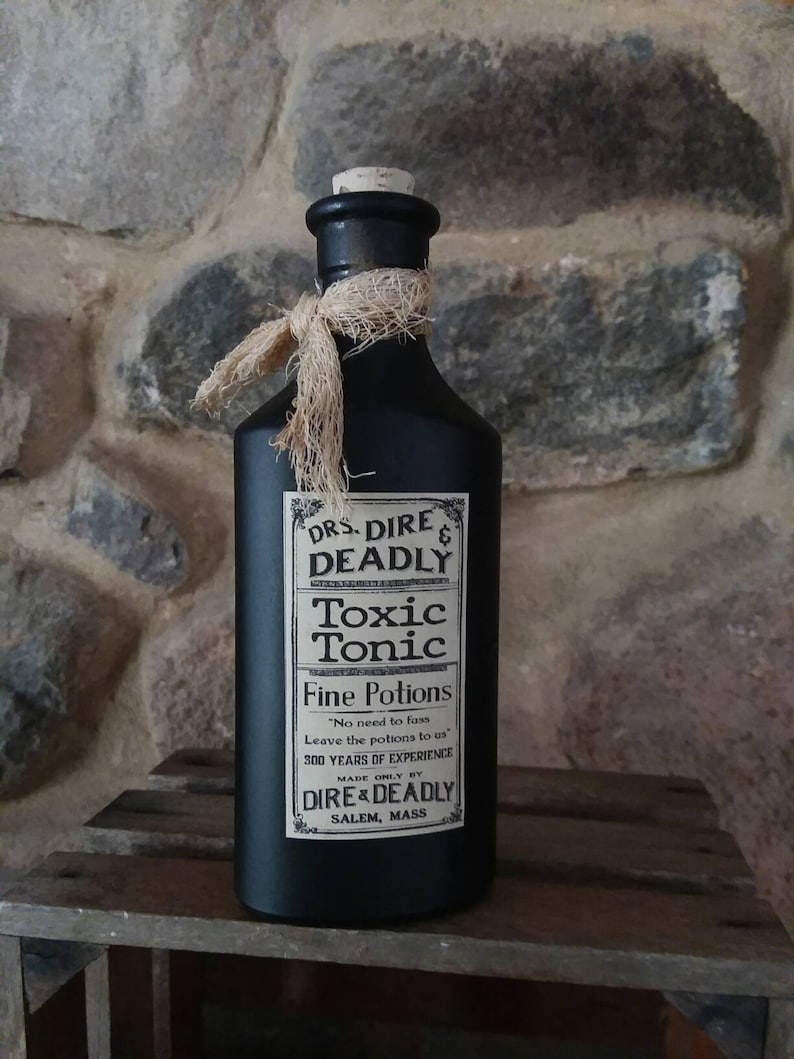 May include: A black glass bottle with a cork stopper and a brown twine bow. The bottle has a label that reads "DRS DIRE & DEADLY Toxic Tonic Fine Potions "No need to fass Leave the potions to us 300 YEARS OF EXPERIENCE MADE ONLY BY DIRE & DEADLY SALEM, MASS"
