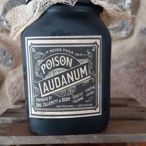 May include: A black glass jar with a white label that reads "IT NEVER FAILS POISON LAUDANUM PREPARED BY DRS. CALAMITY & DOOM SEDATIVE ANODYNE CALMING DEADLY WHITECHAPEL SOUTHWARK". The jar is wrapped in a beige burlap cloth.