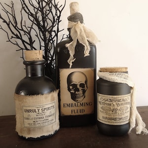 May include: Three black glass bottles with cork stoppers and labels. The labels are printed with vintage-style text and images. The bottles are decorated with burlap and gauze. The labels read "Unruly Spirits", "Embalming Fluid", and "Witch's Powder".