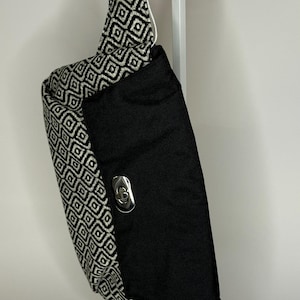 May include: A black and white geometric patterned fanny pack with a black fabric flap and a silver buckle. The fanny pack is hanging on a white display stand.
