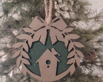 Könnte beinhalten: Ein hölzerner Weihnachtsschmuck mit einem Vogelhaus-Design. Der Schmuck hat eine braune Umrandung und eine grüne Innenseite, verziert mit einer Schleife und Blättern. Er hängt an einer natürlichen Schnur.