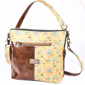 Pode incluir: Bolsa de ombro com estampa de girassóis e painel de couro marrom. A bolsa tem um bolso com zíper e uma alça de couro marrom. A bolsa tem uma pequena etiqueta que diz "Auriety".