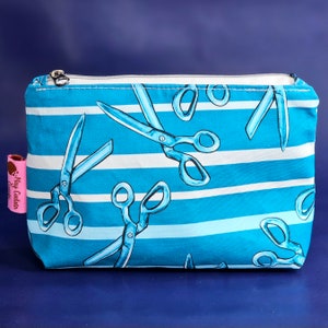 May include: A blue and white striped zippered pouch with a repeating pattern of scissors. The pouch is made of fabric and has a white zipper.
