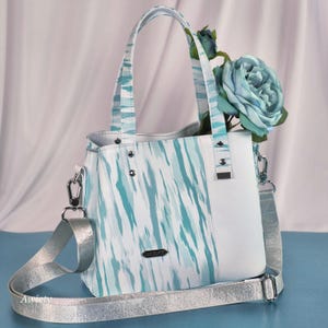 May include: A handbag featuring a white and teal abstract brushstroke pattern, solid white side panels, and silver-toned hardware. It includes two top handles, a detachable silver patterned shoulder strap, and a small metal tag labeled "HANDCRAFTED".