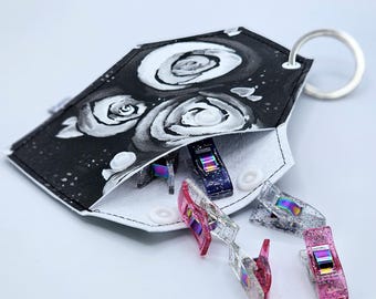 Peut inclure: Une pochette en cuir blanc avec un motif floral noir et blanc et un anneau de clé argenté. La pochette est ouverte et plusieurs clips de couture en plastique irisés en sortent.