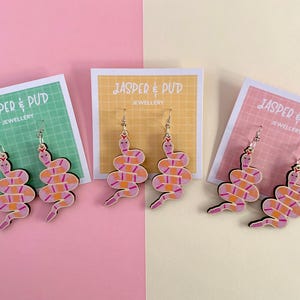 May include: A set of snake-shaped earrings in pink, orange, and white, displayed on cards with the text "JASPER & PUD JEWELLERY". The background is divided into pink, yellow, and green sections.