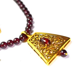 Puede incluir: Un collar con colgante dorado con un diseño de filigrana, con una piedra preciosa roja central y una cuenta a juego. El collar tiene una hebra de pequeñas cuentas redondas de color rojo oscuro. El texto "Victory Is Sweet On Etsy" es visible.