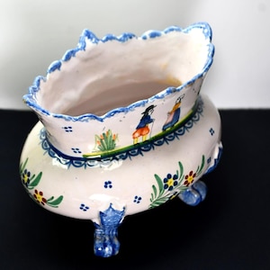 May include: A white ceramic planter with a scalloped rim, adorned with blue trim and floral patterns. The planter features painted figures and rests on three blue feet. A decorative piece.