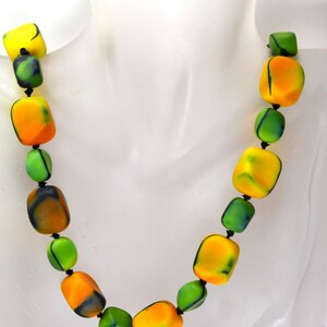 Op de afbeelding: Een kleurrijke ketting met grote, vierkante kralen in geel, oranje en groen. De kralen zijn geregen op een zwart koord, wat een opvallend ontwerp cre&euml;ert. De ketting wordt getoond op een witte mannequin.