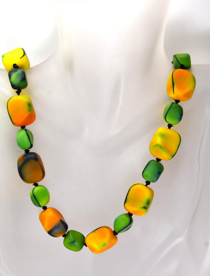 Op de afbeelding: Een kleurrijke ketting met grote, vierkante kralen in geel, oranje en groen. De kralen zijn geregen op een zwart koord, wat een opvallend ontwerp cre&euml;ert. De ketting wordt getoond op een witte mannequin.