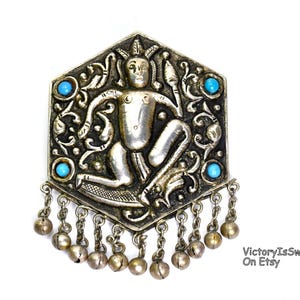 May include: A hexagonal silver-toned brooch featuring a central embossed figure holding a spear, surrounded by intricate scrollwork and four small round turquoise-colored stones. The bottom edge has a row of dangling small metal bells.
