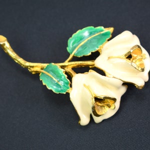 May include: A gold-tone brooch featuring two white enamel roses with green enamel leaves. The roses have a detailed design and are connected by a gold-tone stem.