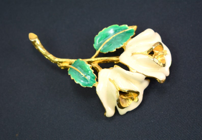 May include: A gold-tone brooch featuring two white enamel roses with green enamel leaves. The roses have a detailed design and are connected by a gold-tone stem.
