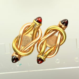 May include: A pair of gold-toned cufflinks with a knot design. Each cufflink features a polished gold finish with a copper-colored center and a red gemstone at each end. The cufflinks are displayed on a light-colored surface.