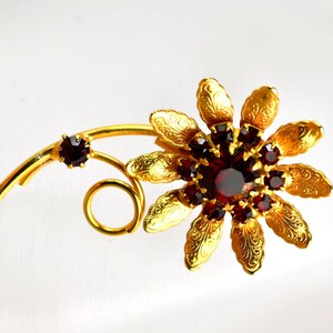 May include: A gold-toned floral brooch with a cluster of red gemstones at the center and on the stem. The brooch features detailed petal and leaf designs, creating a vintage aesthetic. The brooch is a decorative accessory.