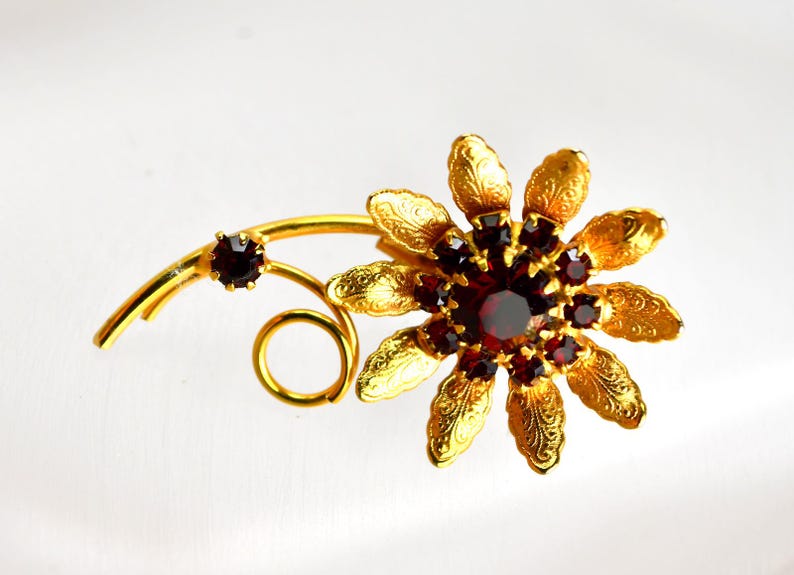 May include: A gold-toned floral brooch with a cluster of red gemstones at the center and on the stem. The brooch features detailed petal and leaf designs, creating a vintage aesthetic. The brooch is a decorative accessory.