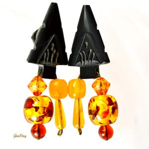 Black carved arrowhead-shaped earrings dangle with faceted orange and amber glass beads. The earrings feature a geometric design and a mix of round, square, and faceted beads in varying shades of orange and amber. The black arrowhead-shaped tops are intricately carved with a repeating pattern.  These statement earrings are perfect for adding a touch of bohemian flair to any outfit.