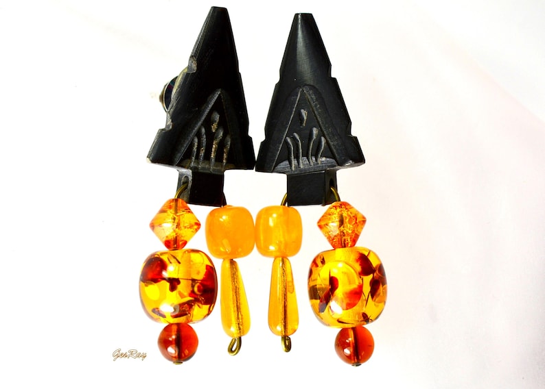 Black carved arrowhead-shaped earrings dangle with faceted orange and amber glass beads. The earrings feature a geometric design and a mix of round, square, and faceted beads in varying shades of orange and amber. The black arrowhead-shaped tops are intricately carved with a repeating pattern.  These statement earrings are perfect for adding a touch of bohemian flair to any outfit.