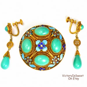 May include: A vintage gold-toned jewellery set comprising a brooch and earrings. The brooch has turquoise stones and floral details. The earrings feature turquoise teardrop pendants. The set is displayed on a white background.