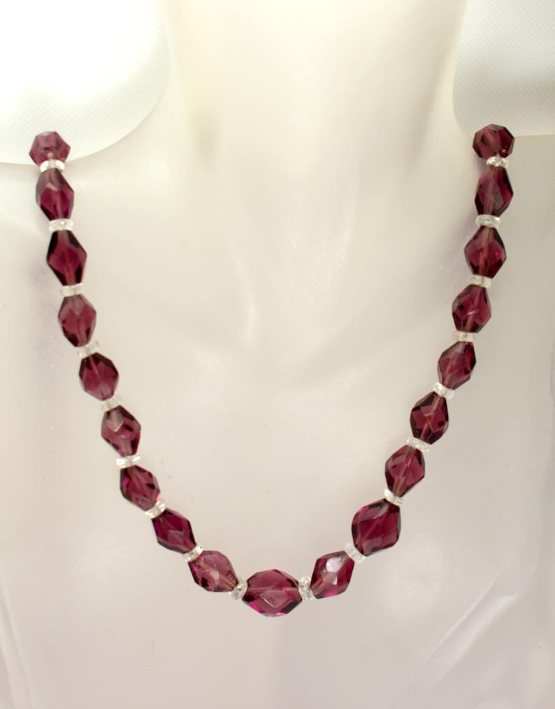 May include: A vintage necklace with faceted, deep purple beads. Small, silver-toned spacers separate the beads. The necklace is displayed on a white mannequin, highlighting its design and colour.