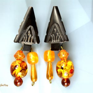 Black carved arrowhead-shaped earrings dangle with faceted orange and amber glass beads. The earrings feature a geometric design and a mix of round, square, and faceted beads in varying shades of orange and amber. The black arrowhead-shaped tops are intricately carved with a repeating pattern.  These statement earrings are perfect for adding a touch of bohemian flair to any outfit.
