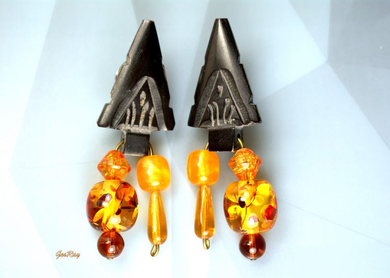Black carved arrowhead-shaped earrings dangle with faceted orange and amber glass beads. The earrings feature a geometric design and a mix of round, square, and faceted beads in varying shades of orange and amber. The black arrowhead-shaped tops are intricately carved with a repeating pattern.  These statement earrings are perfect for adding a touch of bohemian flair to any outfit.