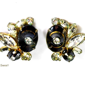 Puede incluir: Un par de pendientes vintage de clip. Cada pendiente presenta una piedra central ovalada negra con un brillante transparente en el centro, rodeada de brillantes transparentes y verde claro. Los pendientes tienen un engaste de metal dorado.