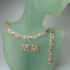 May include: A silver-toned jewellery set comprising a necklace, bracelet, and square earrings. Each piece is embellished with clear, rectangular and square-cut stones. The set is displayed on a light green mannequin head.