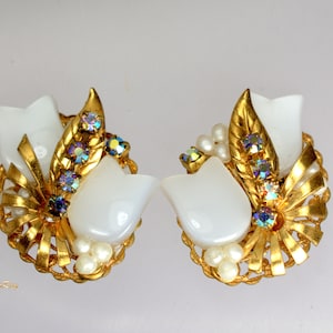 Puede incluir: Un par de pendientes de clip de tono dorado con cabujones blancos en forma de flor, pedrería iridiscente y perlas falsas.