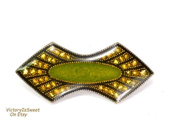 Könnte beinhalten: Vintage Brosche mit geometrischem Design. Die Brosche hat ein ovales Zentrum aus grünem Emaille, das von einem schwarzen Rand umgeben und mit goldfarbenen Akzenten verziert ist. Die Brosche hat eine einzigartige, symmetrische Form.