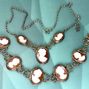 Peut inclure: Un ensemble de bijoux vintage compos&eacute; d'un collier et d'un bracelet. Les deux pi&egrave;ces pr&eacute;sentent des pendentifs cam&eacute;es ovales avec des profils blancs sur fond brun. Le m&eacute;tal dor&eacute; est orn&eacute; de volutes d&eacute;coratives.