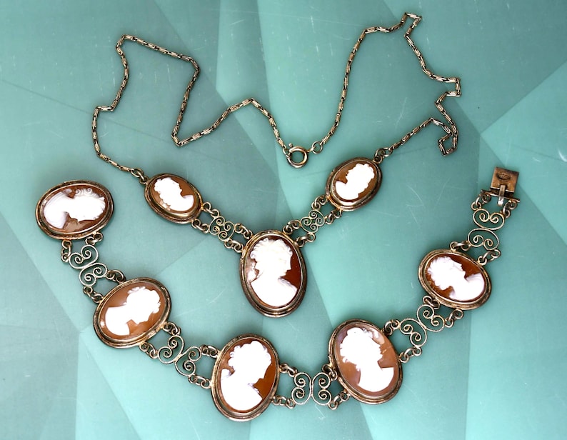 Peut inclure: Un ensemble de bijoux vintage compos&eacute; d'un collier et d'un bracelet. Les deux pi&egrave;ces pr&eacute;sentent des pendentifs cam&eacute;es ovales avec des profils blancs sur fond brun. Le m&eacute;tal dor&eacute; est orn&eacute; de volutes d&eacute;coratives.