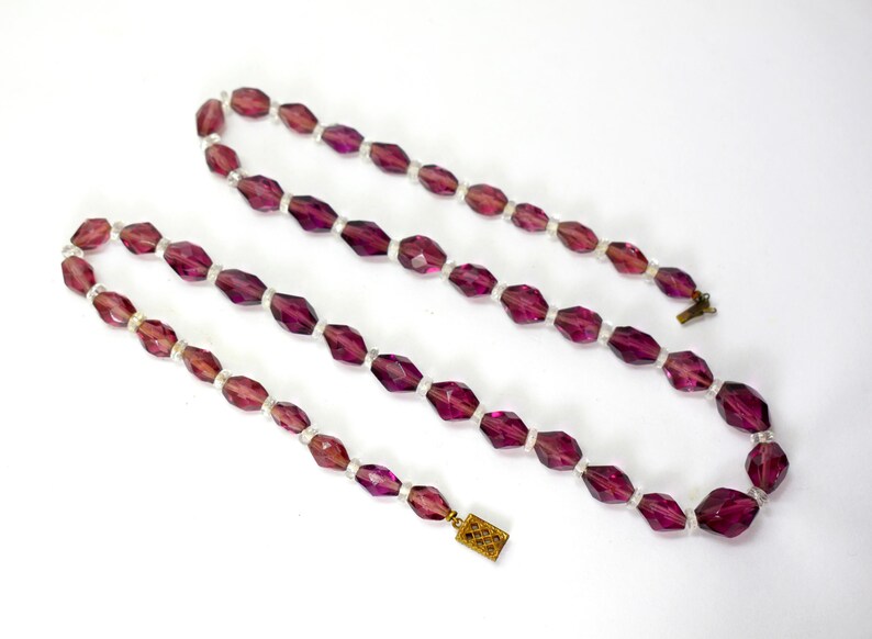 May include: A vintage necklace with faceted, deep purple beads and smaller white beads. The necklace has a gold-toned clasp. The beads are elongated and geometric. The necklace is displayed against a white background.