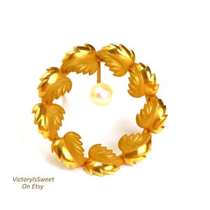 May include: Gold-toned leaf wreath brooch with a dangling pearl. The brooch features a circular design of detailed leaves, surrounding a single, luminous pearl. The jewelry piece is a classic accessory.