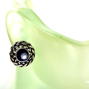 May include: A close-up of a silver-toned stud earring with a woven design around a dark blue center stone. The earring is set against a soft, light green background.