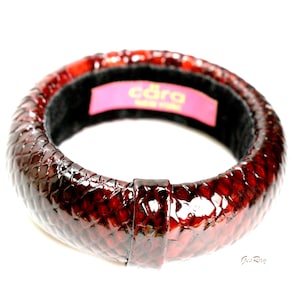 May include: A red, textured bangle bracelet with a glossy finish. The bracelet has a dark interior and a rectangular clasp. The brand name "cara NEW YORK" is visible on a pink label inside the bracelet.