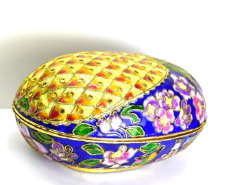 May include: An oval-shaped decorative box with a yellow, diamond-patterned top and a blue base. The box is adorned with gold trim and floral designs in shades of pink, purple, and green. The box is likely made of metal.