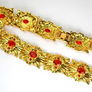 May include: A gold-toned bracelet with ornate floral designs and red gemstone accents. The bracelet features multiple rectangular links with a clasp closure. Small clear crystals are also set into the design.
