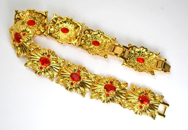 May include: A gold-toned bracelet with ornate floral designs and red gemstone accents. The bracelet features multiple rectangular links with a clasp closure. Small clear crystals are also set into the design.