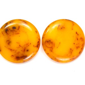 May include: Two circular, smooth, polished amber-colored plugs with a glossy finish. Each piece features a translucent golden-yellow base with dark brown, organic, vein-like inclusions scattered throughout the surface, creating a unique pattern.
