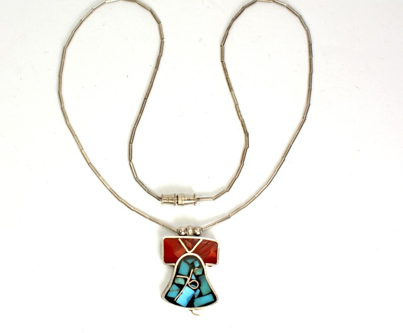 Peut inclure: Un collier en argent avec un pendentif en forme de cloche en turquoise et pierres rouges.