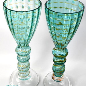May include: Two turquoise-colored stemmed glasses with a clear base. The glasses have a textured pattern with yellow and white accents. The stems are bulbous and the rims are scalloped. The text "GES RAY" is visible at the bottom.