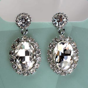 May include: A pair of silver dangle earrings with large oval-shaped clear crystal stones surrounded by smaller clear crystals.