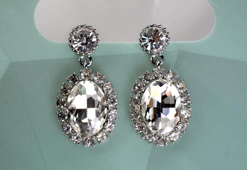 May include: A pair of silver dangle earrings with large oval-shaped clear crystal stones surrounded by smaller clear crystals.