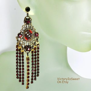 May include: An ornate, gold-toned earring with a cascade of red crystal beads. The earring features a filigree design with a central oval cluster of red crystals and smaller red accents. The earring is shown against a pale green background.