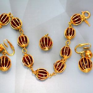 Op de afbeelding: Een gouden armband met een ketting en rode glazen kralen. De kralen zijn in een kooi-achtig ontwerp. Er zijn ook bijpassende oorbellen met rode glazen kralen in een kooi-achtig ontwerp.