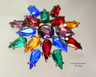 Peut inclure: Broche vintage en forme d'étoile. La broche présente une grosse pierre centrale ronde violette entourée de pierres en forme de goutte dans diverses couleurs, dont le rouge, le bleu, le vert et le jaune. La broche est sertie dans un métal argenté.