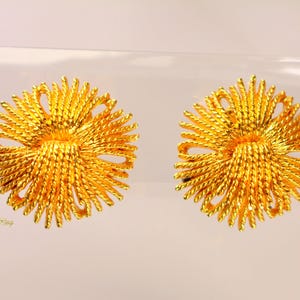 May include: A pair of gold-colored, flower-shaped earrings. Each earring features a central, textured design surrounded by radiating, twisted strands, creating a sunburst effect. The earrings are displayed against a light background.